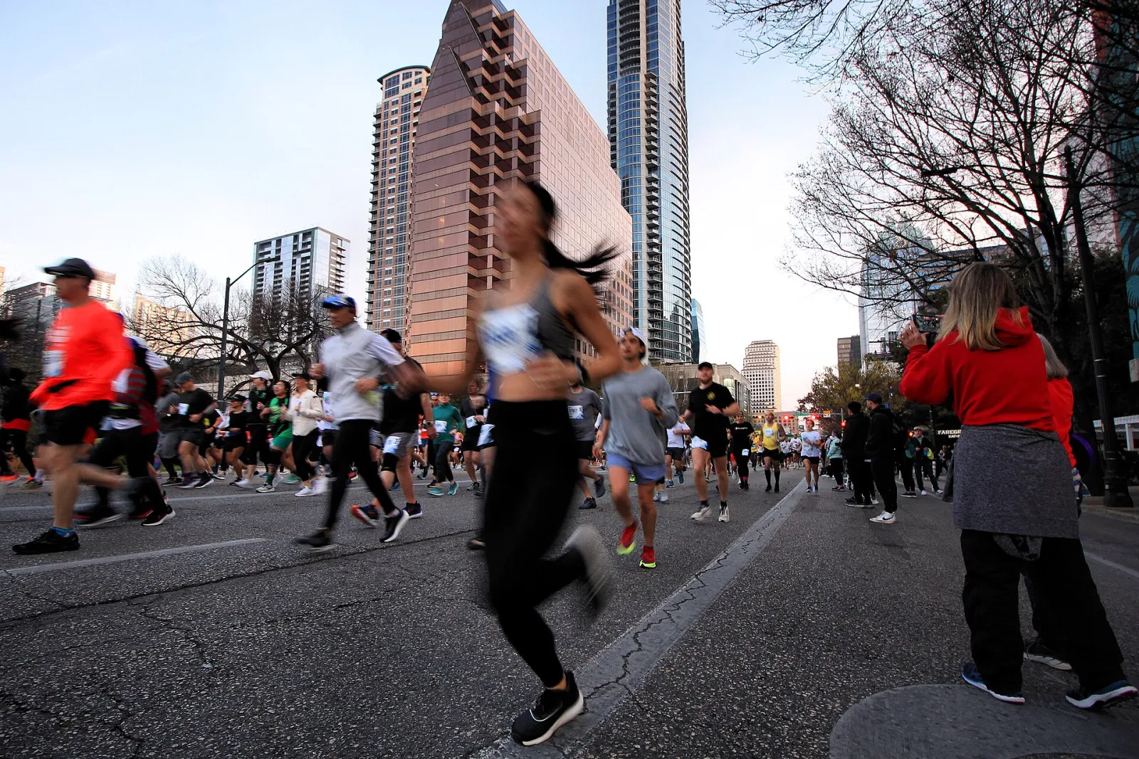 Runners stream down Congress Avenue between downtown Austin high-rises just after the start of the 2023 Austin Marathon, as a spectator takes a photo