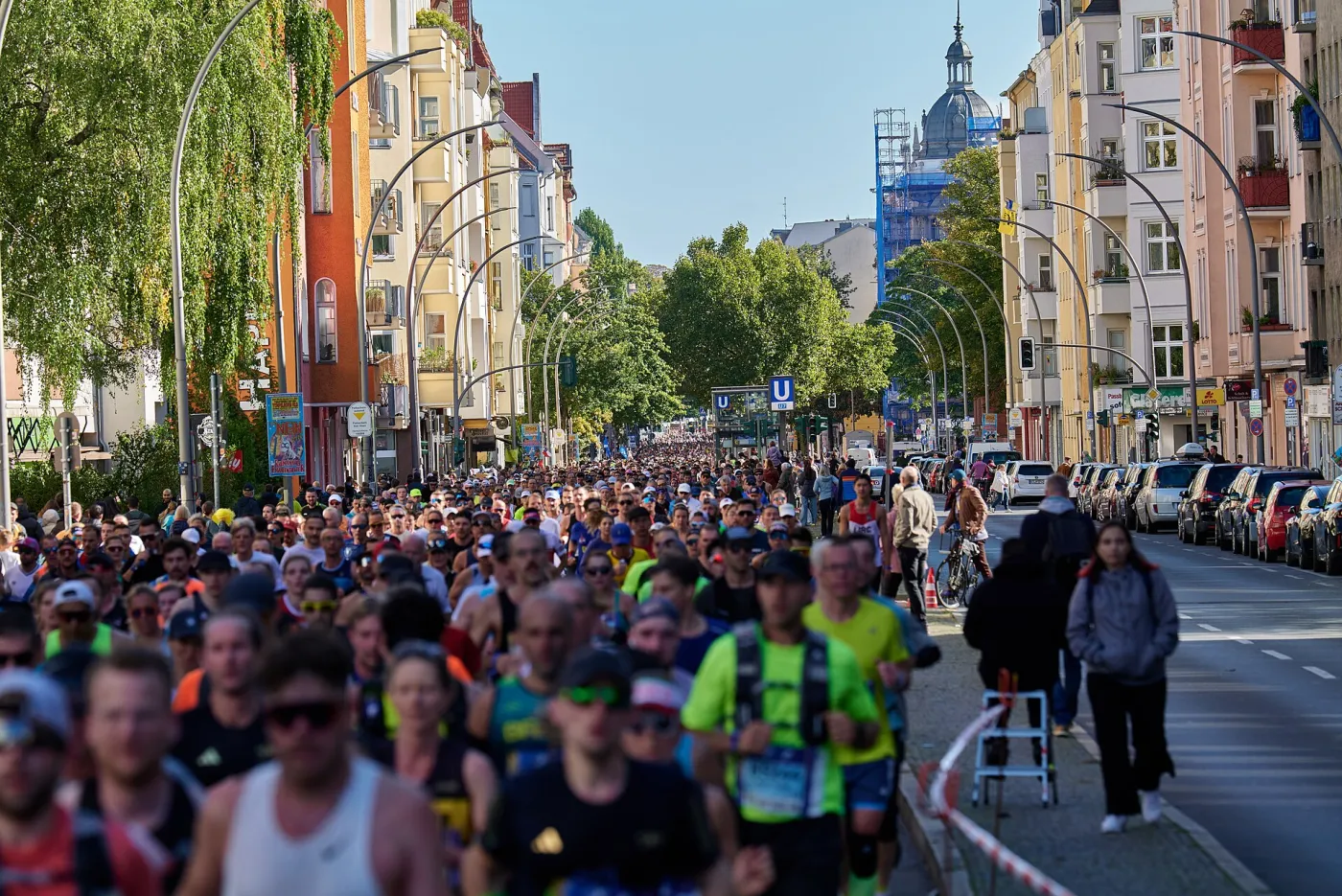 A dense field of Berlin Marathon runners fills Grunewaldstraße in Berlin-Schöneberg, with apartment buildings and a domed tower in the background