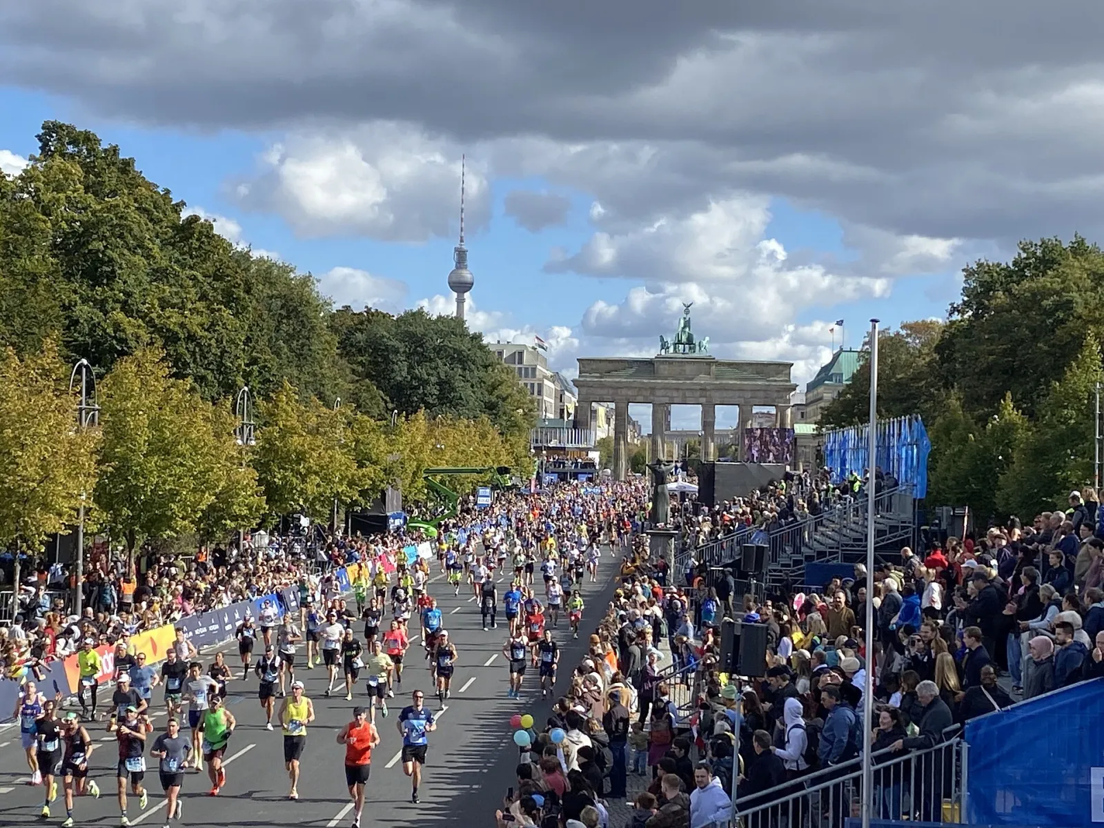 Marathon runners head down a tree-lined avenue toward the Brandenburg Gate finish in Berlin, with grandstands full of spectators on the right