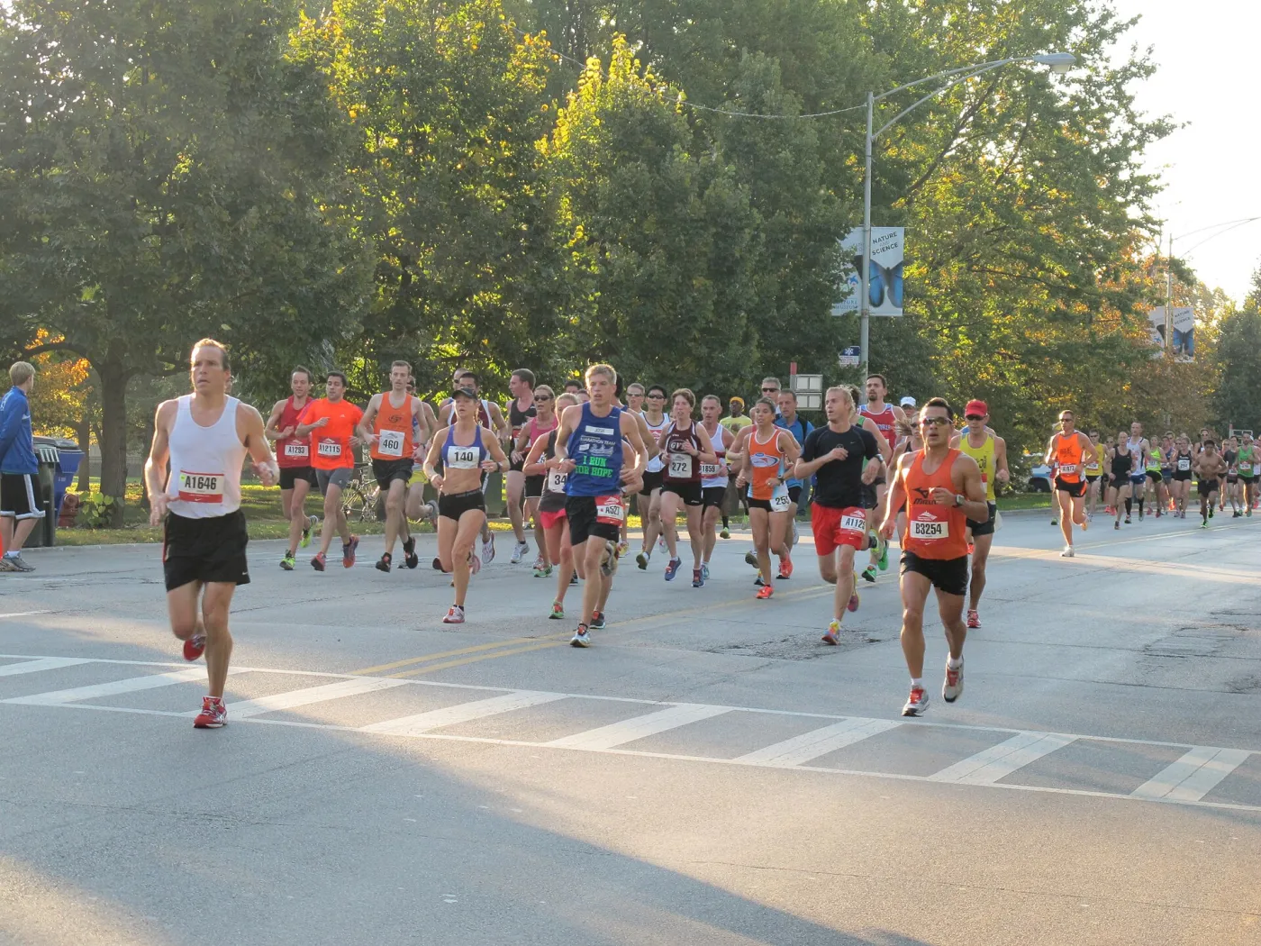 A pack of Chicago Marathon runners with bibs on a tree-lined street, early-morning sun on the course