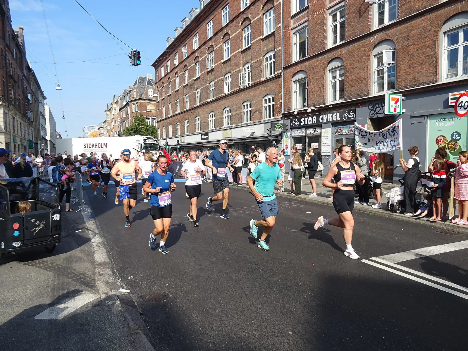 Runners in race bibs pass crowds and brick apartment buildings on Nørrebrogade during the Copenhagen Half Marathon on a sunny day