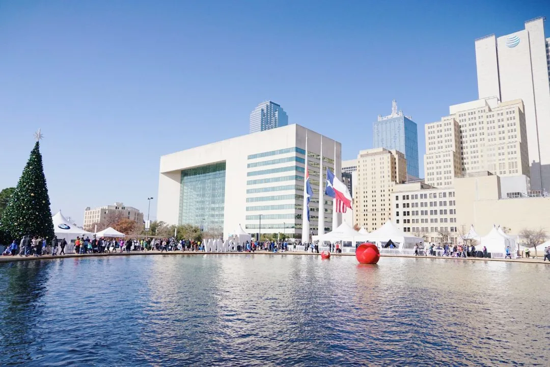 Dallas City Hall Plaza on race weekend, with white tents, a Christmas tree and runners around the reflecting pool under downtown towers