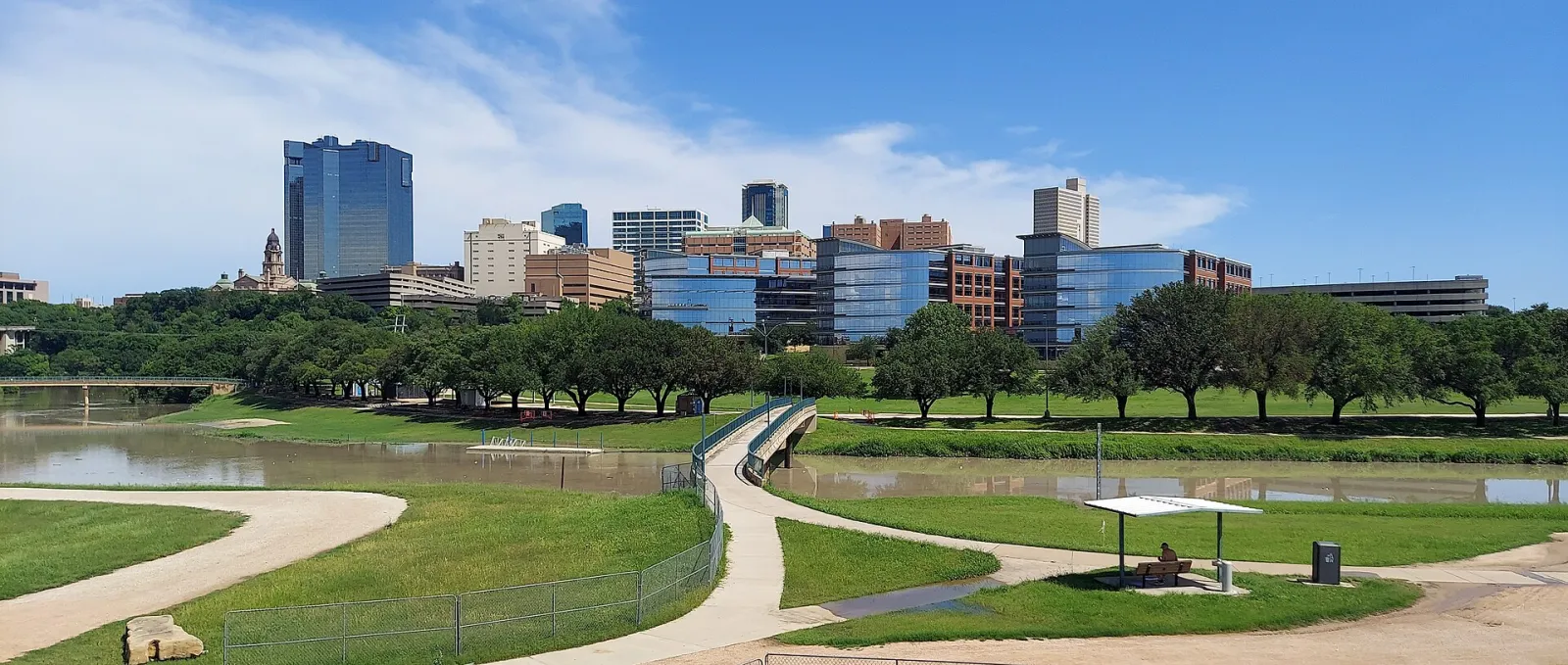 Downtown Fort Worth skyline above the Trinity River, with a paved Trinity Trails path curving across the riverbank