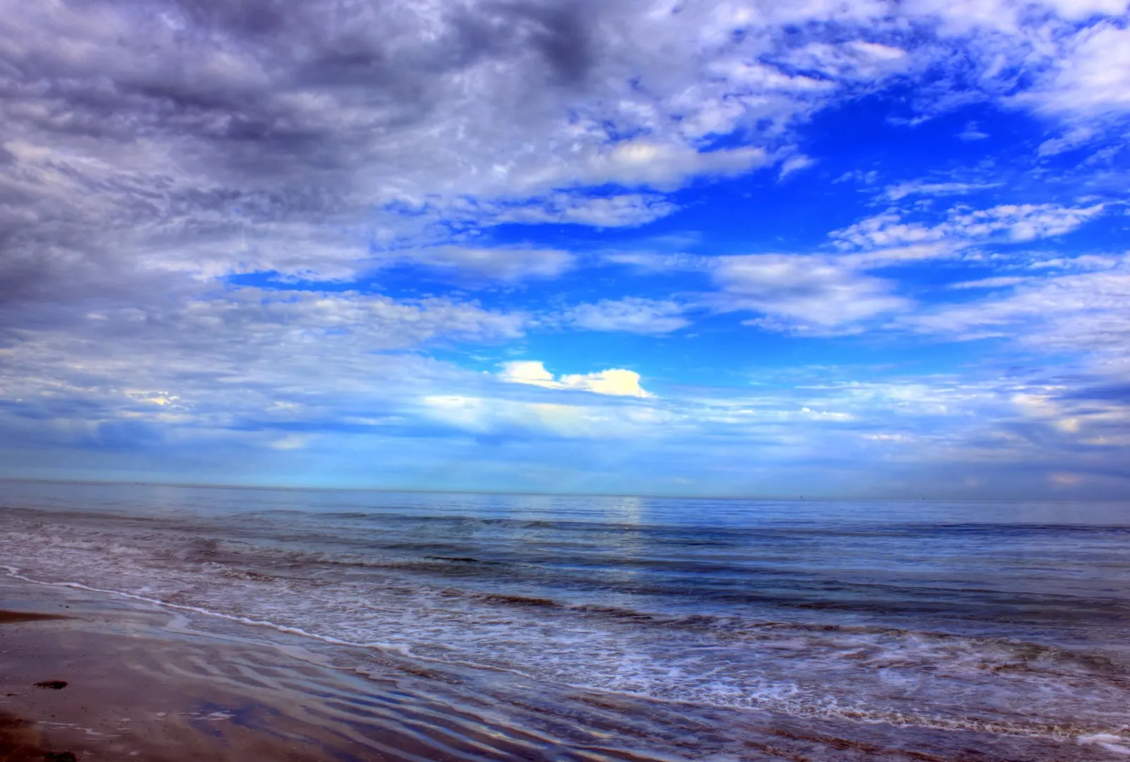 Waves wash up a wide, flat beach on Galveston Island under a sky of broken clouds