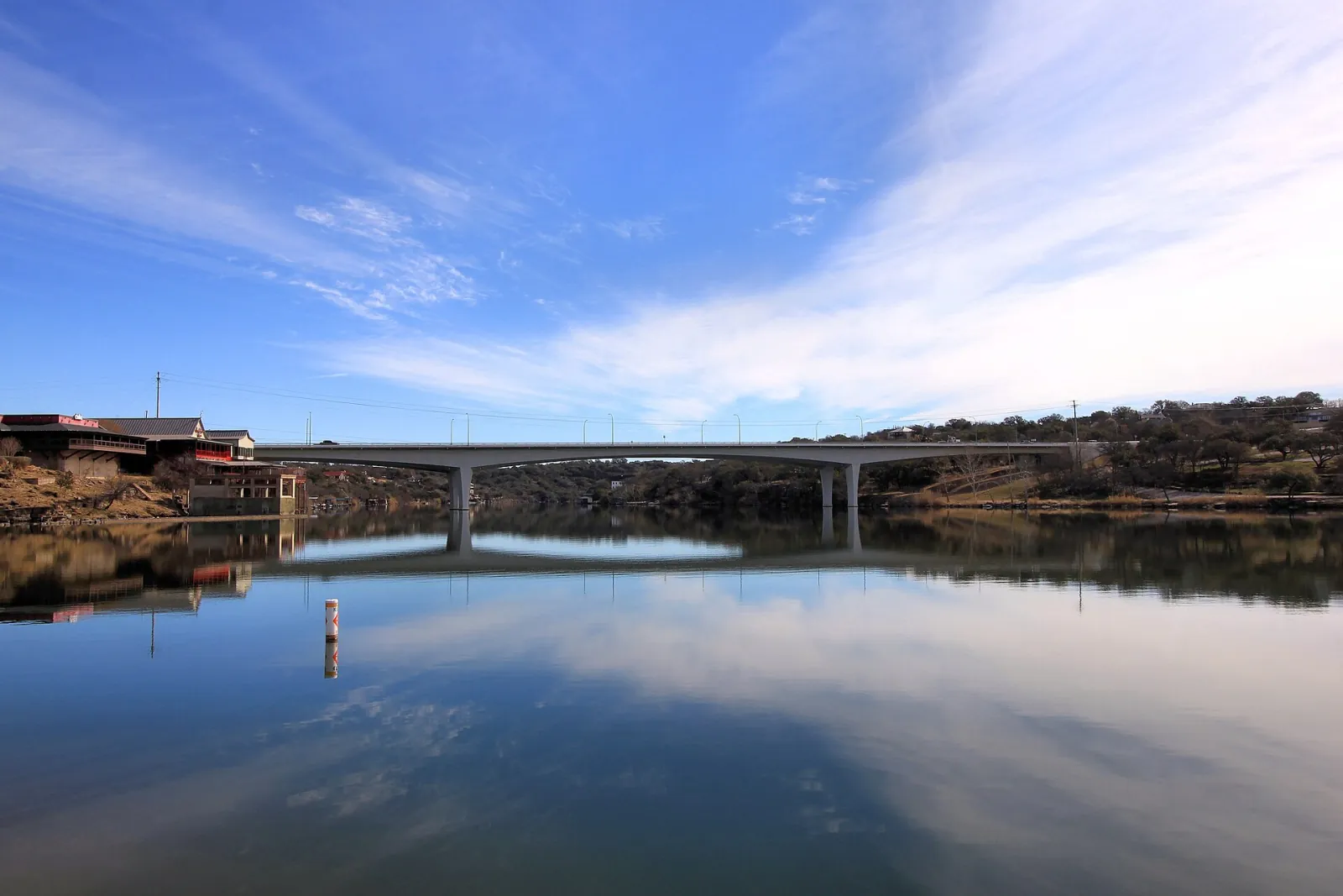 A concrete bridge crosses Lake Marble Falls on a calm day, its reflection mirrored in the water under a blue sky