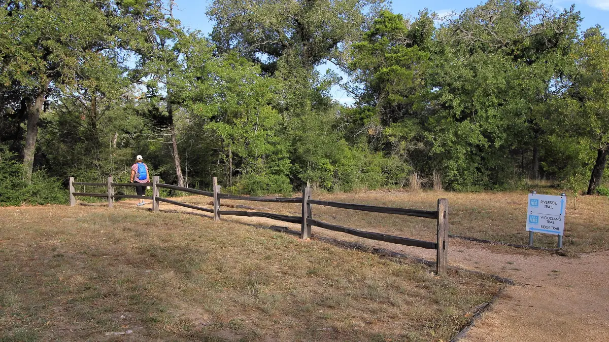 A hiker with a blue pack walking past a split-rail fence onto a dirt trail into oak woods at McKinney Roughs Nature Park