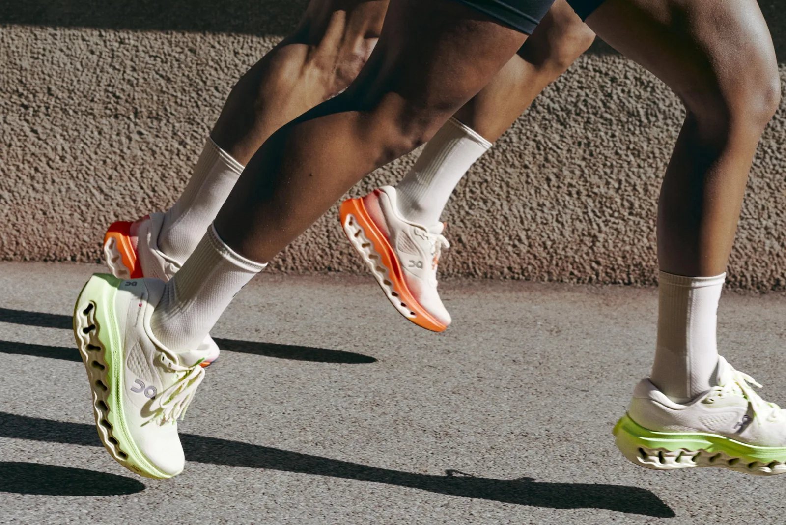 Two runners on a sunny street, one in On Cloudsurfer 3 shoes with lime soles and one in orange, mid-stride