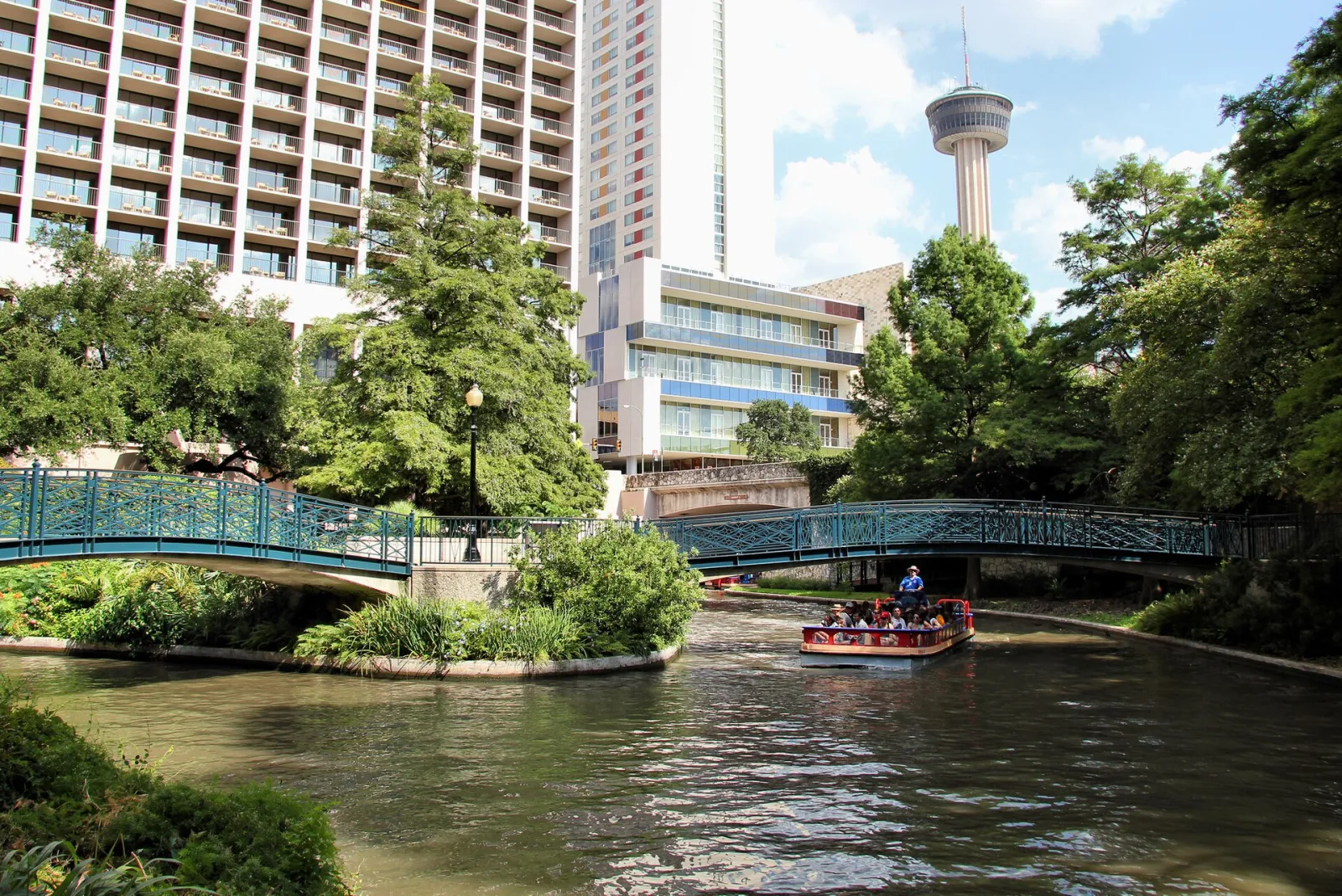 A river barge passes under a teal footbridge on the San Antonio River Walk, with the Tower of the Americas rising behind the trees
