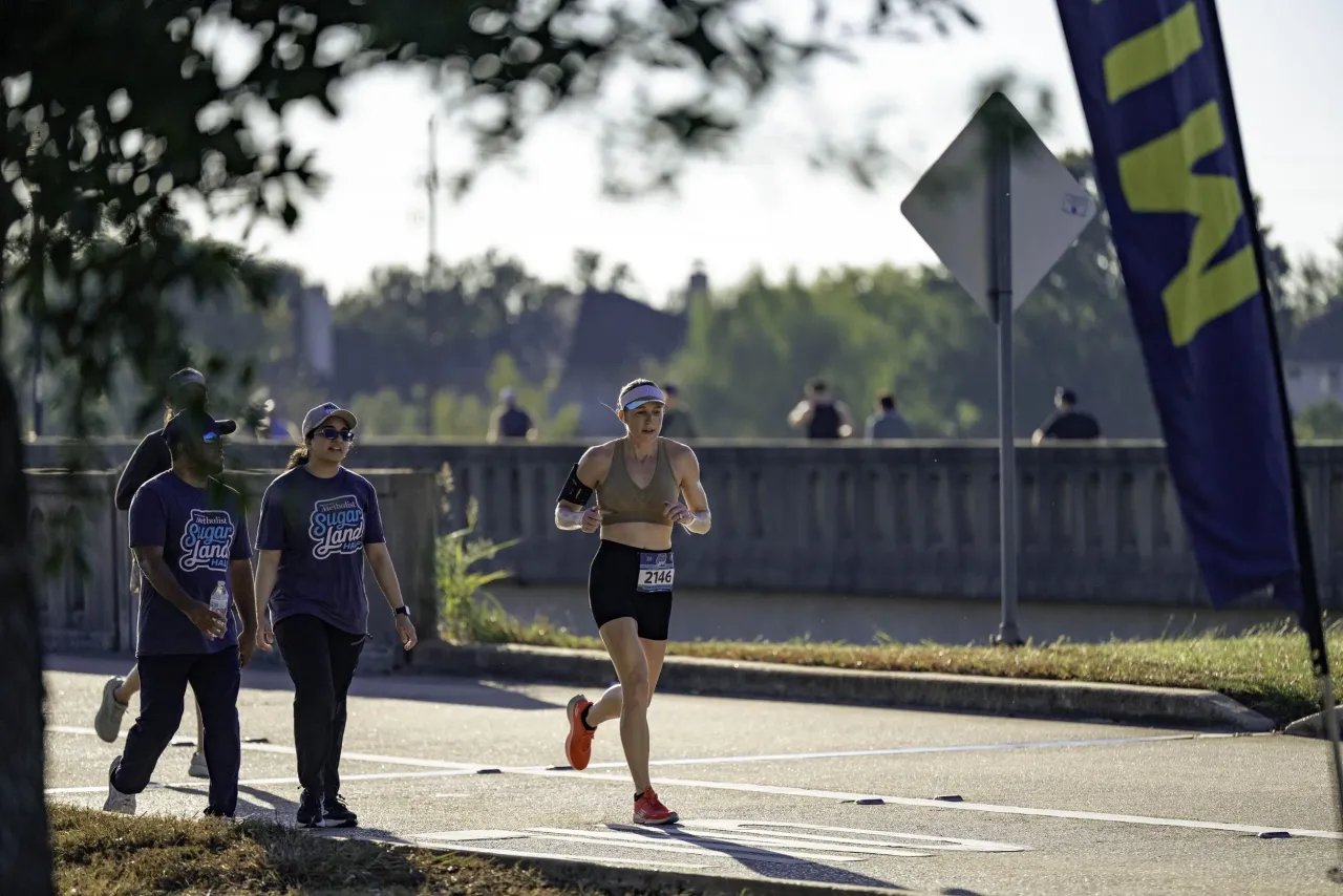 A runner in a visor and black shorts passes volunteers in Sugar Land Half shirts on a bridge road
