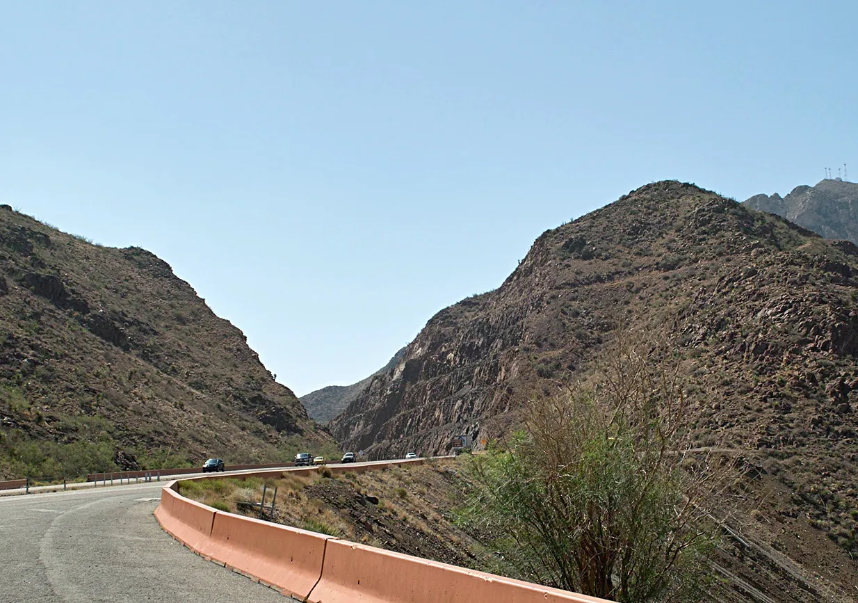 Transmountain Road curving through a cut in the Franklin Mountains in El Paso, with rocky brown slopes on both sides and a clear blue sky